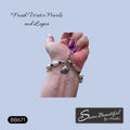 A bracelet displayed on a person's wrist, featuring white freshwater pearls, dark blue beads, and a stainless steel silver-toned charm, set against a light blue background with text overlays.