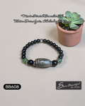 Black beaded bracelet with a central stone on a light surface next to a small potted plant, with branding visible.