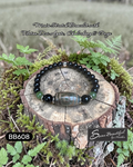 Black beaded bracelet with central stone on a wooden stump surrounded by greenery, with 'Sseau Beautiful' branding.