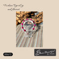 A bracelet displayed on an acrylic stand.  Bracelet is made with bright fuchsia tiger eye, garnet, and silver-tone stainless steel beads, on a textured beige background with wood and a striped fabric.
