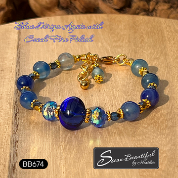 A beaded bracelet with blue agate beads, a large central deep blue bead, small iridescent blue-and-gold decorative beads, and gold-tone spacer rings and clasp hardware, displayed on a wooden surface. Text overlays read 'Blue Stripe Agate with Czech Fire Polish' and 'BB674'; a logo at bottom right reads 'Sseau Beautiful by Heather'.
