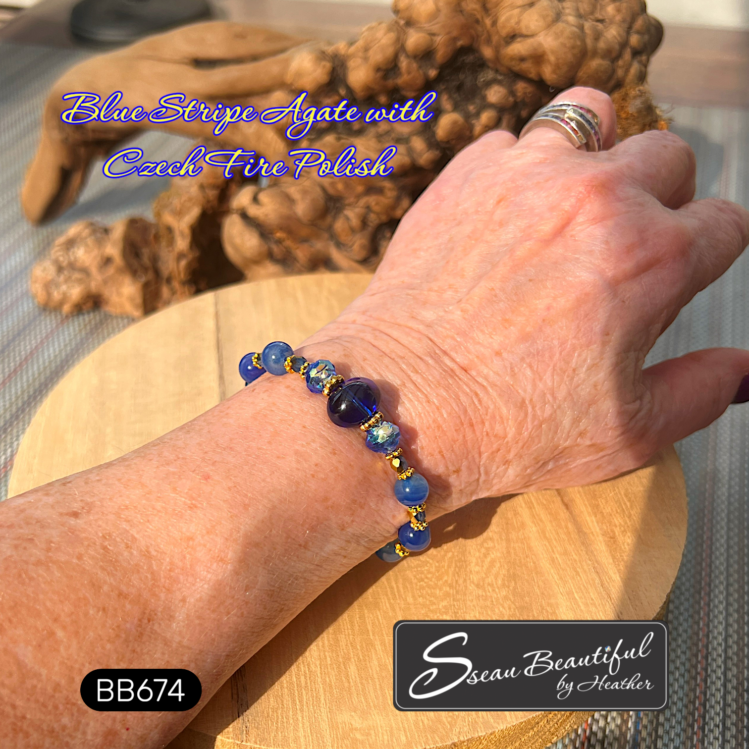 A close-up of a person's wrist wearing a beaded bracelet made of blue agate and Czech fire polish beads with gold spacer accents, resting on a wooden stand with decorative wood in the background. Text overlays read 'Blue Stripe Agate with Czech Fire Polish', 'BB674', and 'Sseau Beautiful by Heather'.