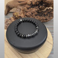 A men's gemstone stretch bracelet rotating on a black display stand against a rustic driftwood background. The bracelet features glossy black onyx beads, polished silver-tone stainless steel disc accents, two metallic grey hematite rounds, and a detailed carved hematite lion head focal bead. The video transitions with a spin to a black and white circular logo for Sseau Beautiful by Heather, displaying the website sseaubeautiful.com. Handcrafted in Coquitlam, BC.