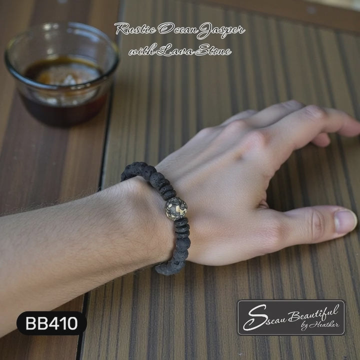 Man's stretch bracelet made with rustic Ocean Jasper and Lava on a man's wrist resting on a wood table with maker's "Sseau Beautiful" logo in the lower right corner.
