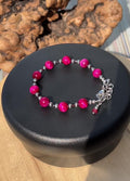 A bracelet with bright fuchsia tiger's eye, garnet, and silver-tone stainless steel beads and extender on a black rotating display.