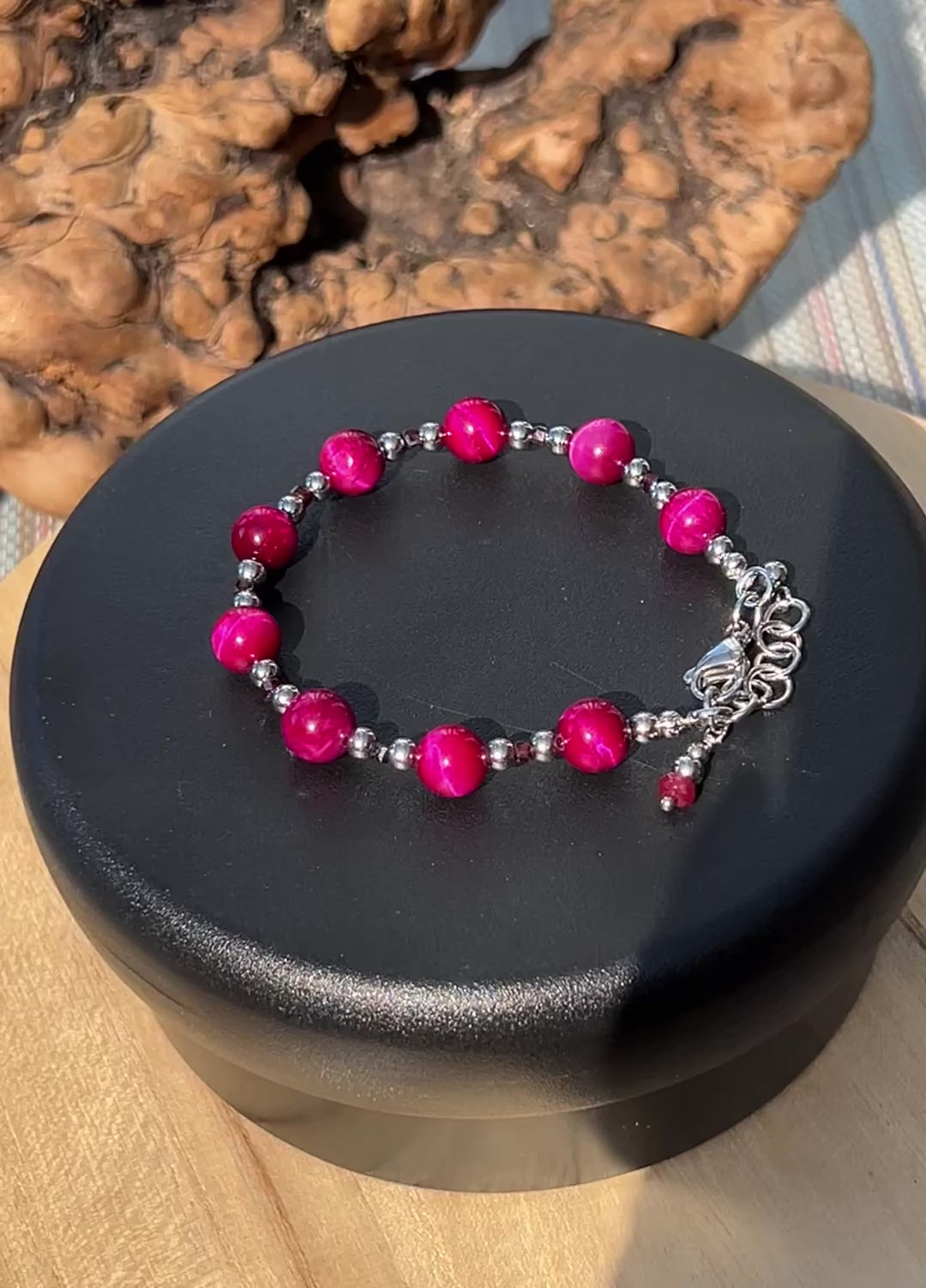 A bracelet with bright fuchsia tiger's eye, garnet, and silver-tone stainless steel beads and extender on a black rotating display.