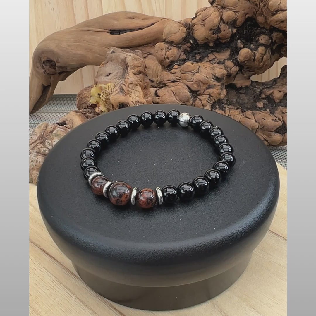 A 5-second product video showcasing the BB612 Men's Gemstone Stretch Bracelet. The video features the bracelet smoothly rotating on a black display stand against a light wood background with natural driftwood accents. The design features glossy black onyx beads, tarnish-resistant stainless steel spacers, and a center focal of brick-red and black swirled Mahogany Obsidian beads. The video concludes with a clean black-and-white Sseau Beautiful by Heather brand logo outro.