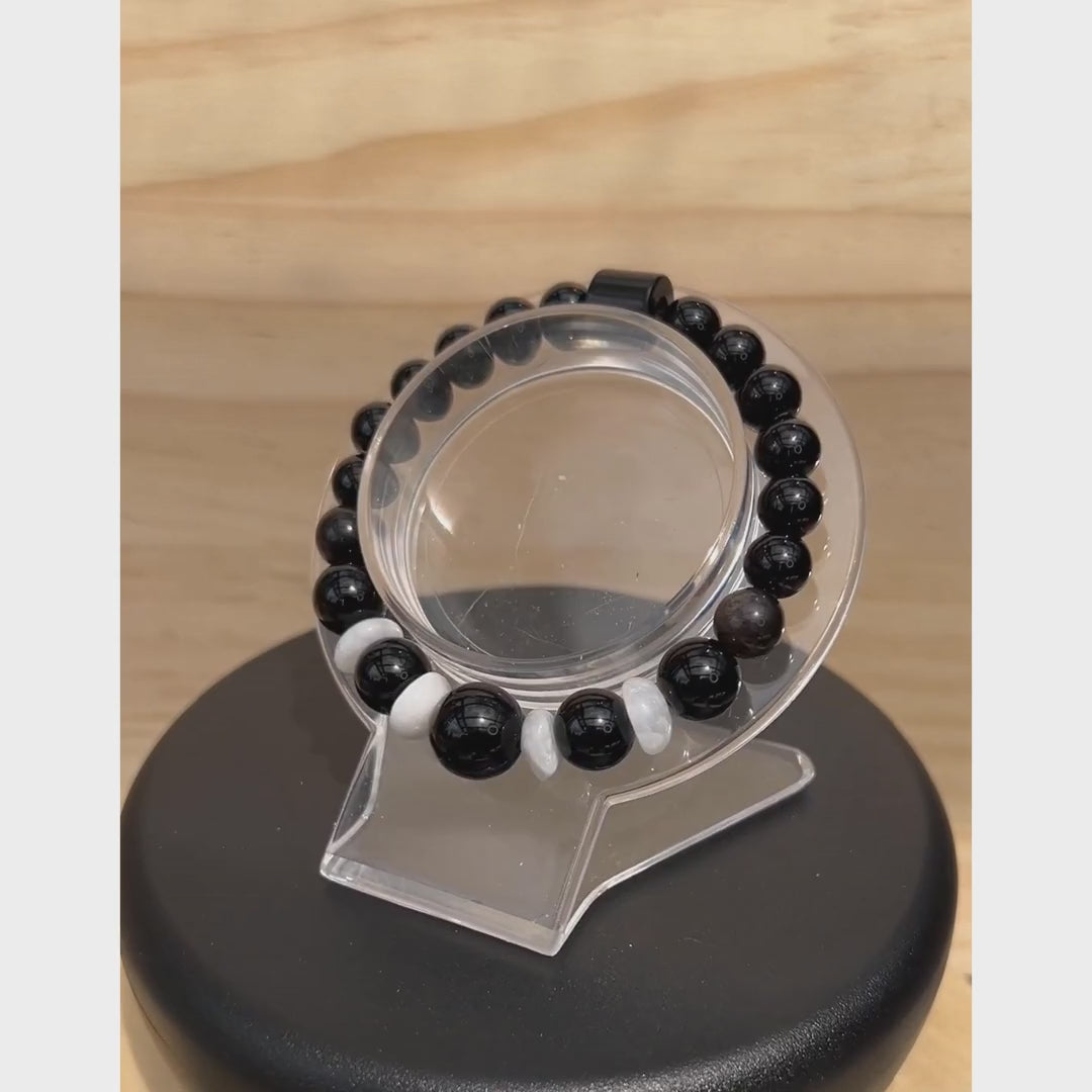 A 360-degree rotating video of a handcrafted unisex gemstone stretch bracelet displayed on a clear stand. The 6-inch bracelet features a large, high-shine 12mm black onyx center bead flanked by white moonstone discs, polished black onyx beads, and a silver obsidian tube bead. Handcrafted by Sseau Beautiful in Coquitlam, BC.